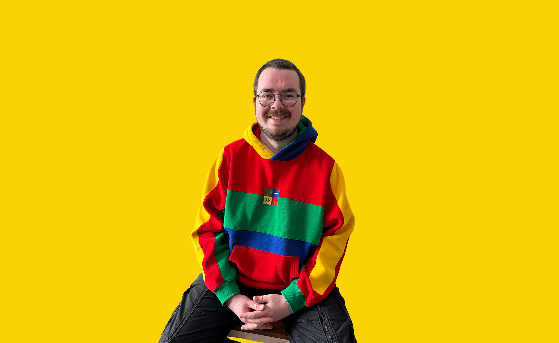 Managing Director, Miles Cant, sat down on a chair wearing a red, green and yellow jumper from cool.shirtz. Featured on a solid yellow background.